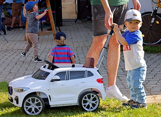 Drei Jungen spielen mit einem Spielzeugauto in einem Park. Ein Junge läuft, während ein anderer im Auto sitzt und von einem Erwachsenen geschoben wird. Ein Fahrrad ist dahinter geparkt.
