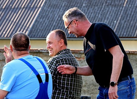 Drei Männer stehen vor einem Haus, einer von ihnen trägt eine Brille und eine Uhr. Der Mann in der Mitte lächelt und scheint zu sprechen. Der Mann auf der rechten Seite zeigt auf den Mann auf der linken Seite.