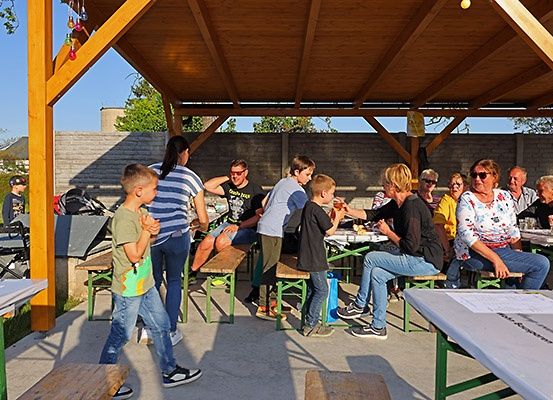 Eine Gruppe von Menschen sitzt und steht um Tische herum unter einem hölzernen Pavillon, isst und unterhält sich. Einige tragen Sonnenbrillen. Der Bereich ist im Freien.