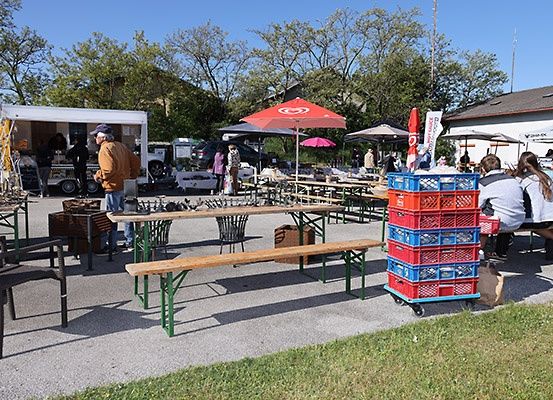 Ein Outdoor-Marktaufbau mit Tischen, Bänken und Sonnenschirmen. Ein Mann steht neben einem Food Truck. Ein Gebäude mit einer roten Flagge ist im Hintergrund sichtbar.