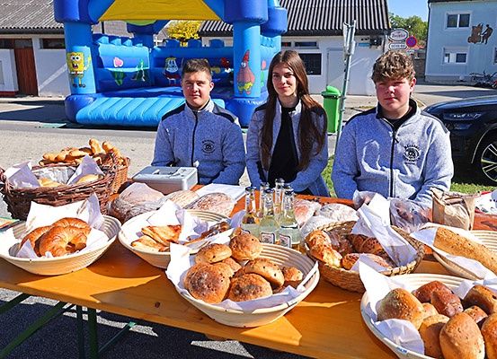 Drei Personen stehen hinter einem Tisch voller verschiedener Brote und Gebäckstücke, mit einem bunten aufblasbaren Hüpfburg im Hintergrund.