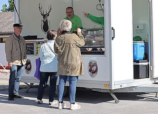 Drei Personen stehen vor einem weißen Food-Truck mit einem aufgehängten Hirschkopf. Ein Mann macht ein Foto, während zwei Frauen hineinschauen.