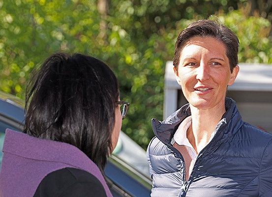 Zwei Frauen stehen vor einem Auto, eine lächelt und spricht mit der anderen, die eine Brille trägt.