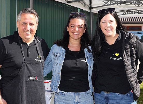 Drei Personen, zwei Frauen und ein Mann, lächeln für ein Foto in einer sonnigen Außenumgebung, wahrscheinlich bei einem Grill-Event.