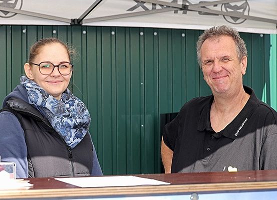 Zwei Personen stehen lächelnd hinter einem Tisch in einer Kabine, die Frau trägt eine Brille und einen Schal, der Mann ein schwarzes Hemd.
