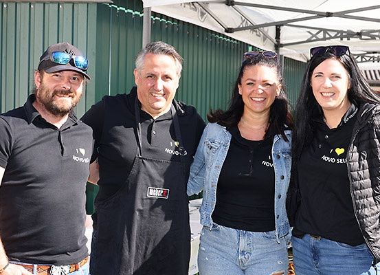 Vier Menschen lächeln für ein Foto, zwei Frauen und zwei Männer, alle in schwarzen T-Shirts. Sie stehen unter einem Zelt, wahrscheinlich bei einer Essensveranstaltung.