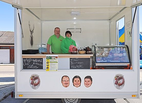 Zwei Personen in grünen Uniformen stehen lächelnd in einem Food-Truck. Der Truck ist mit Logos und einer Kreidetafel-Menü dekoriert. In der Nähe befindet sich ein Haus mit einem Reh-Silhouette an der Wand.