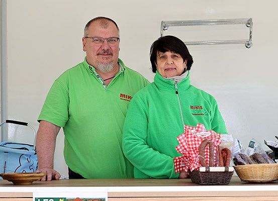 Zwei Personen stehen hinter einem Tresen, tragen grüne Uniformen und lächeln. Sie sind umgeben von Körben mit Lebensmitteln, einem Handtuch und einer Tasche. Ein silberner Ständer ist an der Wand hinter ihnen befestigt.