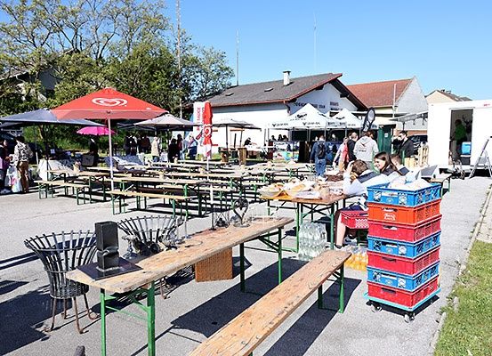 Ein Freiluftbereich mit mehreren Tischen mit Bänken und Menschen, die herumlaufen. Mehrere Zelte sind ebenfalls aufgebaut. Vor den Zelten steht ein Tisch mit Gegenständen darauf, und eine Frau sitzt neben einem Stapel Kisten.