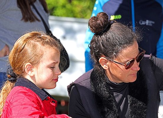 Ein junges Mädchen in einer roten Jacke spricht mit einer Frau mit Sonnenbrille und einer mit Fell gefütterten Jacke. Sie stehen im Freien.