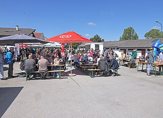 Eine Outdoor-Versammlung mit Menschen, die unter Schirmen auf Bänken sitzen und einige stehen, mit Gebäuden und Bäumen im Hintergrund.