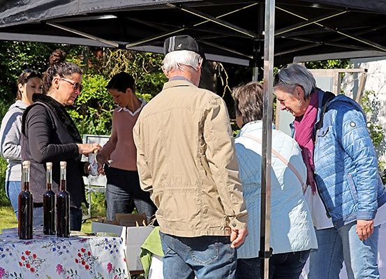 Eine Gruppe von Menschen versammelt sich unter einem Baldachin, möglicherweise auf einem Markt. Flaschen mit Wein stehen auf dem Tisch, und einige schenken Wein in Gläser ein.
