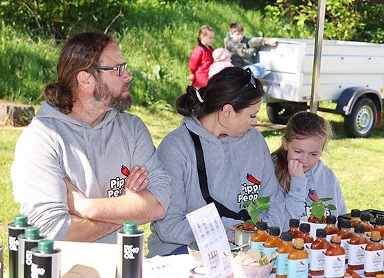 Drei Personen stehen an einem Stand mit Flaschen Soße. Ein Mann hat die Arme verschränkt. Im Hintergrund sitzen zwei Kinder und ein Anhänger.
