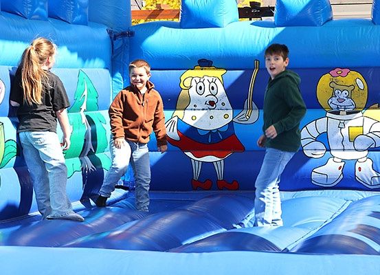 Drei Kinder spielen in einem blauen aufblasbaren Sprunghaus mit Cartoon-Charakteren. Die Kinder lächeln und scheinen Spaß zu haben.