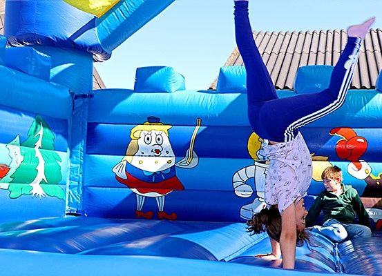Ein junges Mädchen balanciert auf den Händen in einem blauen aufblasbaren Sprunghaus mit Comicfiguren, während ein Junge am Boden zuschaut.