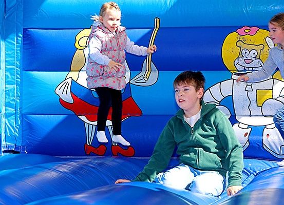 Drei Kinder spielen in einem blauen aufblasbaren Sprunghaus mit Cartoon-Charakteren. Ein Mädchen springt, ein Junge sitzt und ein weiteres Mädchen steht daneben.