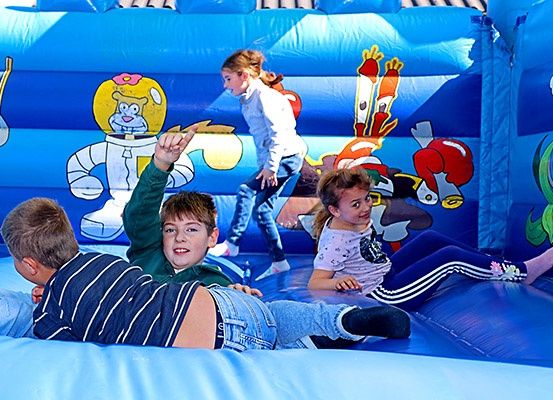 Mehrere Kinder spielen in einem blauen aufblasbaren Sprunghaus mit Comic-Zeichenfiguren an den Wänden. Ein Kind liegt auf der Rutsche, während ein anderes in der Nähe steht.