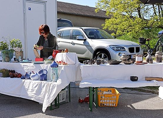 Eine Frau ordnet Gegenstände auf einem Tisch im Freien an, in der Nähe eines Autos und Fahrräder. Zwei Tische mit weißen Tüchern zeigen verschiedene Objekte, darunter Vasen und Teekannen.