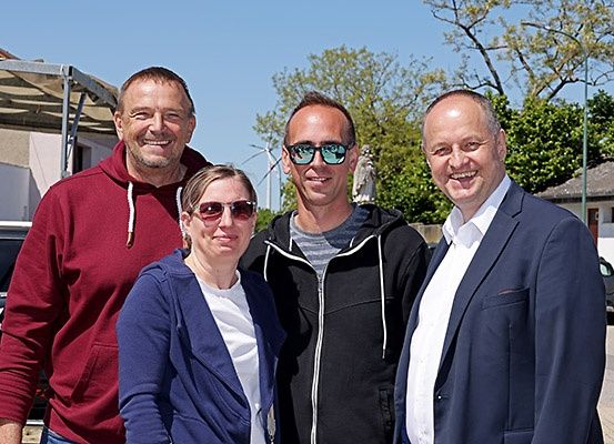 Vier Menschen lächeln vor einem Gebäude mit Vordach und Bäumen. Ein Auto ist in der Nähe geparkt.