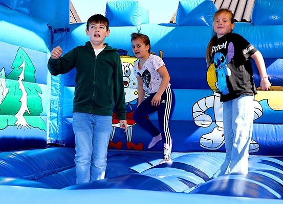 Drei Kinder springen in einem blauen aufblasbaren Sprunghaus, das mit Zeichentrickfiguren verziert ist. Der Junge links trägt eine grüne Jacke, das Mädchen in der Mitte ein weißes Shirt und das Mädchen rechts ein schwarzes Shirt mit einer Zeichentrickfigur.