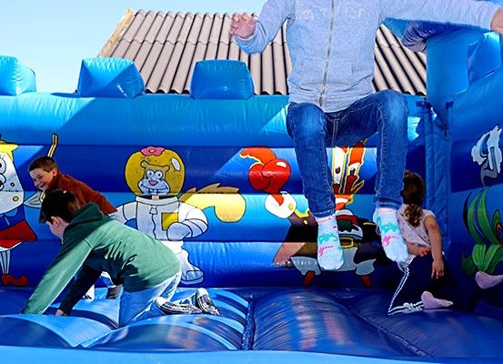 Ein Kind springt in einem blauen aufblasbaren Sprunghaus mit Cartoon-Charakteren. In der Nähe spielen zwei Kinder auf dem Aufblasbaren.