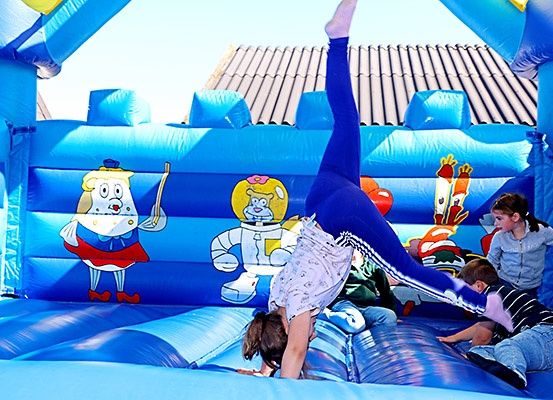 Ein Mädchen macht einen Handstand in einem blauen aufblasbaren Hüpfhaus mit Cartoon-Charakteren an der Seite. Zwei Kinder spielen auf dem Boden darin.