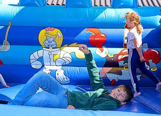 Drei Kinder spielen auf einem blauen aufblasbaren Hüpfhaus mit Comic-Zeichen. Ein Junge liegt, während ein Mädchen steht und ein anderes in der Nähe spielt.