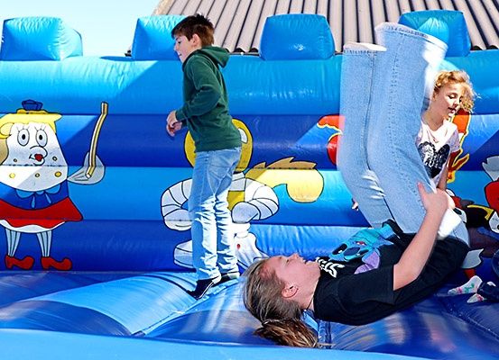 Drei Kinder spielen auf einem blauen aufblasbaren Sprunghaus. Ein Kind steht, während ein anderes auf dem Rücken fällt. Ein drittes Kind steht in der Nähe.