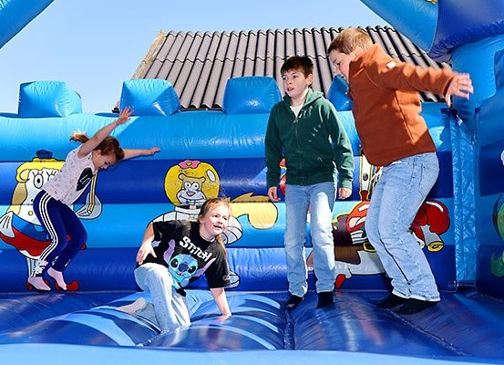 Vier Kinder spielen in einem blauen aufblasbaren Hüpfburg mit Comicfiguren. Ein Mädchen springt, ein anderes Mädchen hockt und zwei Jungen stehen in der Nähe.