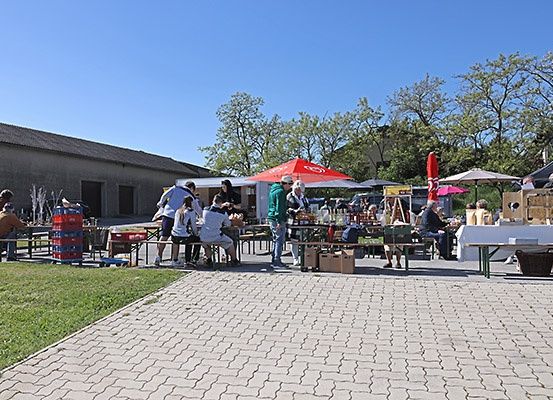 Im Freien liegende Markt mit Tischen, Sonnenschirmen und Körben. Menschen sitzen unter Schirmen und um Tische herum. Ein Gebäude ist im Hintergrund.