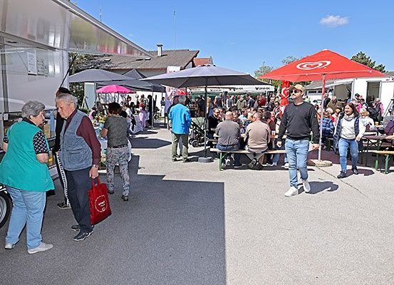 Ein Freiluftmarkt mit mehreren Imbisswagen, Sonnenschirmen und Menschen, die an Tischen sitzen, einige mit Kinderwagen, unter blauem Himmel.