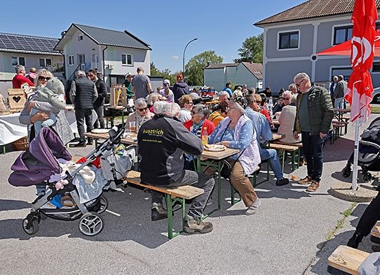 Eine Outdoor-Versammlung mit Menschen, die an Tischen sitzen, einige mit Kinderwagen, unter Schirmen in einem sonnigen Außenbereich.