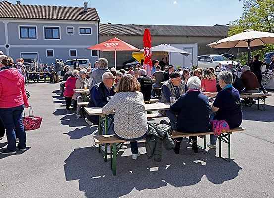 Eine Gruppe älterer Menschen trifft sich zu einer Mahlzeit vor einem Gebäude. Sie sitzen an Tischen unter Sonnenschirmen. Eine Frau geht mit einem Korb vorbei.