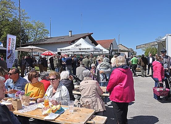 Eine Gruppe von Menschen versammelt sich für eine Veranstaltung im Freien, sitzt um Picknicktische herum mit Essen. Im Hintergrund sind Zelte und Gebäude aufgebaut, und ein Banner mit roten Schriftzeichen ist sichtbar.
