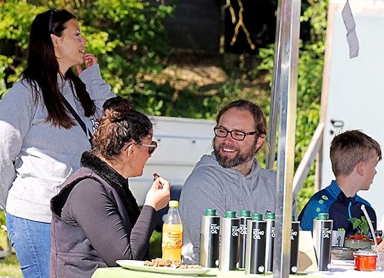 Eine Familie genießt ein Essen im Freien. Eine Frau mit Brille isst, während ein Mann lächelt. Ein Junge schaut sie an. Es gibt einen Tisch mit Essen, Getränken und Pflanzen.