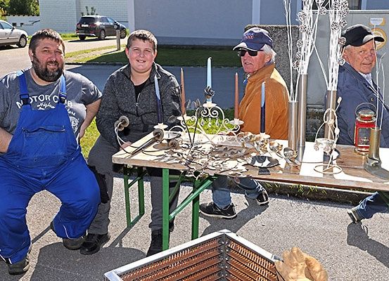 Vier Männer sitzen an einem Tisch mit verschiedenen Metallarbeiten darauf. Sie scheinen an den Kunstwerken zu arbeiten.