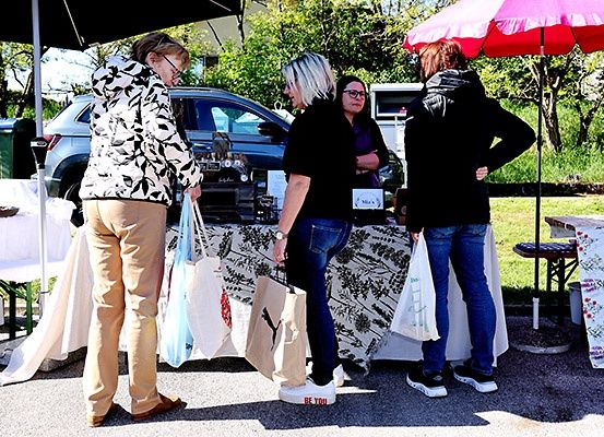 Drei Frauen stehen auf einem Freiluftmarkt, eine hält eine Puma-Tasche, eine andere eine weiße Tasche und eine dritte eine blaue Tasche. Sie stehen unter einem Baldachin, hinter ihnen ist ein schwarzes Auto geparkt.