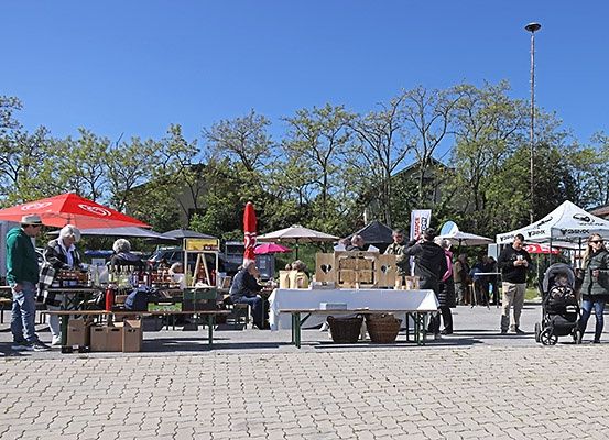 Ein Outdoor-Markt mit verschiedenen Ständen unter Zelten. Einige Leute sitzen, andere stehen und stöbern. Ein Kinderwagen und ein Kinderwagen mit einem Kind sind zu sehen.