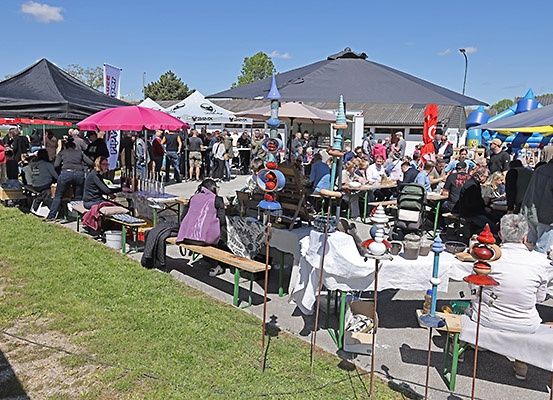 Eine große Menschenmenge versammelt sich unter Zelten, Fahnen und Regenschirmen für einen Markt im Freien. Tische und Bänke sind mit verschiedenen ausgestellten Artikeln aufgestellt, und Menschen stöbern und interagieren.