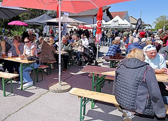 Eine Gruppe von Menschen versammelt sich bei einer Veranstaltung im Freien, sitzt um Tische unter Schirmen, einige tragen Sonnenbrillen und Mützen. Der Hintergrund deutet auf ein Festival hin, mit mehreren Zelten und Bannern.