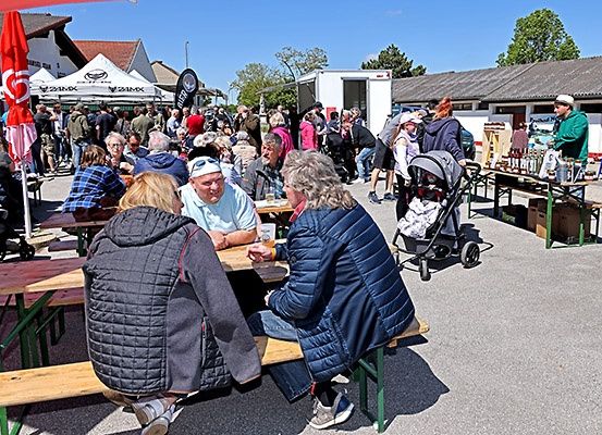 Eine Menschenmenge versammelt sich bei einer Outdoor-Veranstaltung. Einige sitzen an Tischen, während andere stehen oder herumlaufen. Ein Kinderwagen ist in der Nähe, und es gibt Zelte und einen Imbisswagen im Hintergrund.