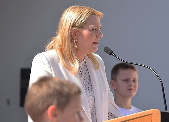 Eine Frau mit blonden Haaren, die einen weißen Blazer trägt, spricht in ein Mikrofon an einem Rednerpult, wobei zwei Jungen sie ansehen.