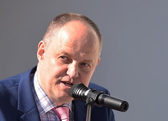 Ein Mann in einem blauen Anzug und einer rosa Krawatte spricht in ein Mikrofon, möglicherweise bei einer Veranstaltung.