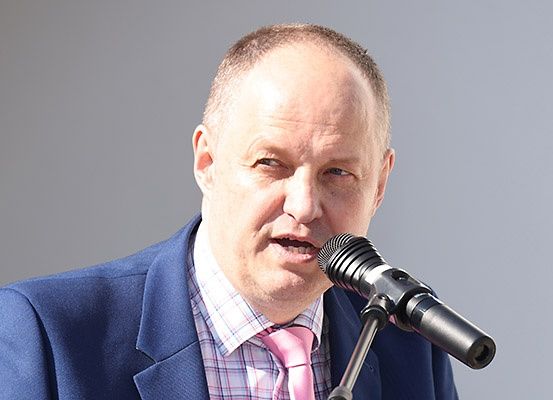Ein Mann in einem blauen Anzug und einer rosa Krawatte spricht in ein Mikrofon, wahrscheinlich bei einer Veranstaltung.