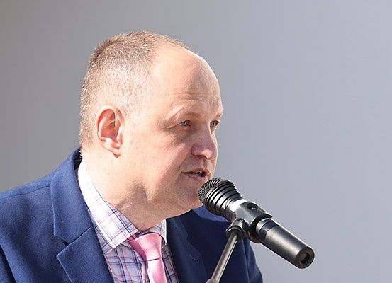 Ein Mann in einem blauen Anzug und einer rosa Krawatte spricht in ein Mikrofon. Er wirkt konzentriert und ernst.