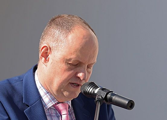 Ein Mann in formeller Kleidung spricht in ein Mikrofon, seine Augen geschlossen. Das Mikrofon ist schwarz mit einem Ständer.