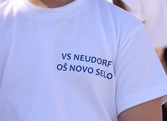Eine Nahaufnahme eines weißen T-Shirts mit blauem Text, der 'VS NEUDORF OS NOVO SELO' auf der Vorderseite steht.