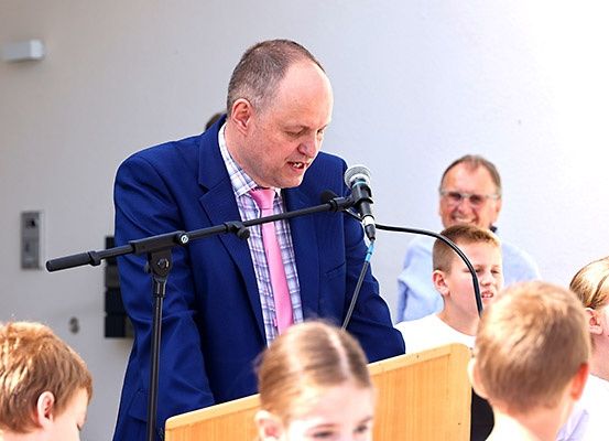 Ein Mann in einem blauen Anzug und einer rosa Krawatte spricht in ein Mikrofon an einem Rednerpult, während Kinder und ein anderer Mann zuhören.