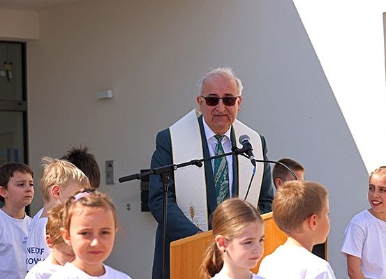 Ein älterer Mann steht an einem Rednerpult und hält eine Rede vor einer Gruppe von Kindern in weißen T-Shirts, die vor einem Gebäude mit großen Fenstern stehen.