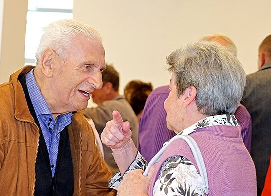 Ein älterer Mann und eine ältere Frau unterhalten sich lebhaft und stehen in einem Raum, in dem mehrere Personen im Hintergrund zu sehen sind.